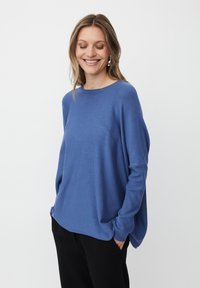 Frau lächelt mit Händen in der Tasche, trägt einen blauen Langarmpullover und schwarze Hosen vor einem schlichten weißen Hintergrund.