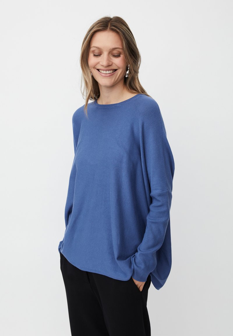 Frau lächelt mit Händen in der Tasche, trägt einen blauen Langarmpullover und schwarze Hosen vor einem schlichten weißen Hintergrund.