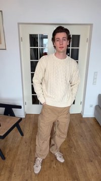 Młody mężczyzna stoi w pomieszczeniu, ubrany w kremowy sweter warkoczowy, beżowe spodnie typu cargo i jasne sportowe buty, ręce w kieszeniach.