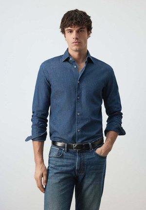 Junger Mann mit lockigem Haar, der ein blaues Jeanshemd und Jeans trägt, mit einer Hand in der Tasche, steht vor einem schlichten hellen Hintergrund.