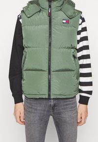 Zelená péřová vesta s vysokým límcem, předním zipem a cvoky; na hrudi je značková nášivka a postranní kapsy; nošena přes pruhované tričko.