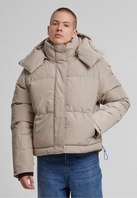 Beige puffert jacket magas gallérral, cipzáras zár�ódással és hímzett logóval. Enyhén fényes textúrával rendelkezik, az ujjaknál pedig elasztikus részletekkel.