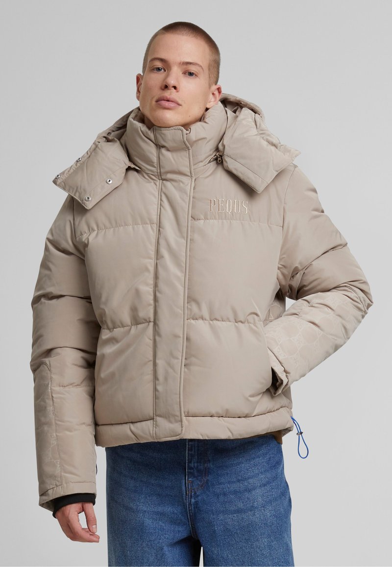 Beige puffert jacket magas gallérral, cipzáras záródással és hímzett logóval. Enyhén fényes textúrával rendelkezik, az ujjaknál pedig elasztikus részletekkel.