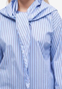 Chemise bleu clair avec fines rayures verticales blanches et rouges, stylisée avec un tissu rayé assorti drapé autour du cou.