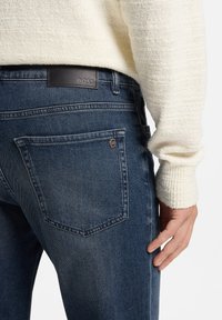 Vaqueros de mezclilla azul oscuro con una etiqueta de cuero BOSS en la cintura, que presentan un diseño clásico de cinco bolsillos y un sutil desgaste en la tela.