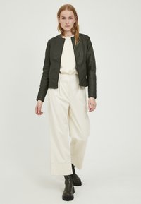 Giacca bomber in pelle nera con collo rotondo, indossata sopra un top crema e pantaloni bianchi larghi; il modello si trova su uno sfondo chiaro.
