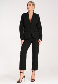 Czarna marynarka dopasowana z klapami, zapięciem na jeden guzik i dwiema przednimi kieszeniami; w połączeniu z czarnymi, dopasowanymi spodniami i odkrytymi czółenkami.