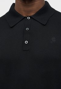 Gros plan sur un polo noir avec un col côtelé, trois boutons noirs, et un logo brodé discret sur la poitrine.