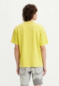 Yellow cotton t-shirt with a crew neck and short sleeves, viewed from the back, paired with light gray denim shorts featuring a faded design.