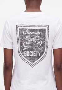 Personne portant un T-shirt blanc avec un motif de bouclier noir dans le dos, comportant les mots Samsøe Society et un graphique de ruban.