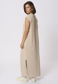 Beige ärmelloses Maxikleid mit seitlichen Schlitzen, aus weichem Stoff, mit lockerer Passform. Kombiniert mit weißen klobigen Sneakers.