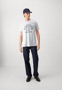 Tricou alb din bumbac cu grafică "HILFIGER NEW YORK", asociat cu blugi din denim închis cu cusături contrastante și pantofi din piele neagră.