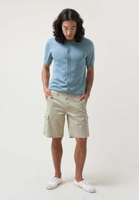 Cárdigan de punto azul claro de manga corta con botones, combinado con pantalones cortos de cargo beige. Modelo lleva zapatos de lona blancos.