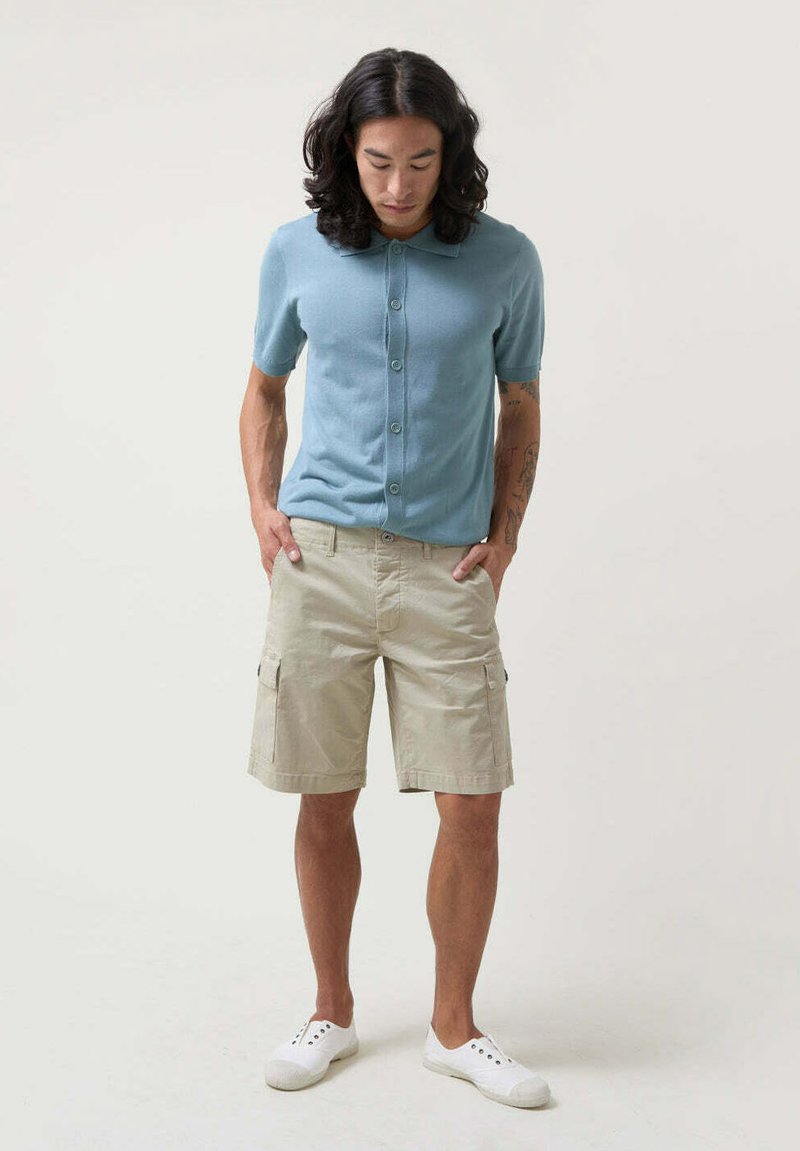 Cárdigan de punto azul claro de manga corta con botones, combinado con pantalones cortos de cargo beige. Modelo lleva zapatos de lona blancos.