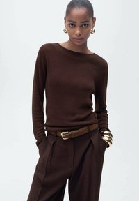 Pull en maille marron avec un col rond, associé à un pantalon marron taille haute et ajusté, et une ceinture dorée. Gros bracelet jonc en or en accent.
