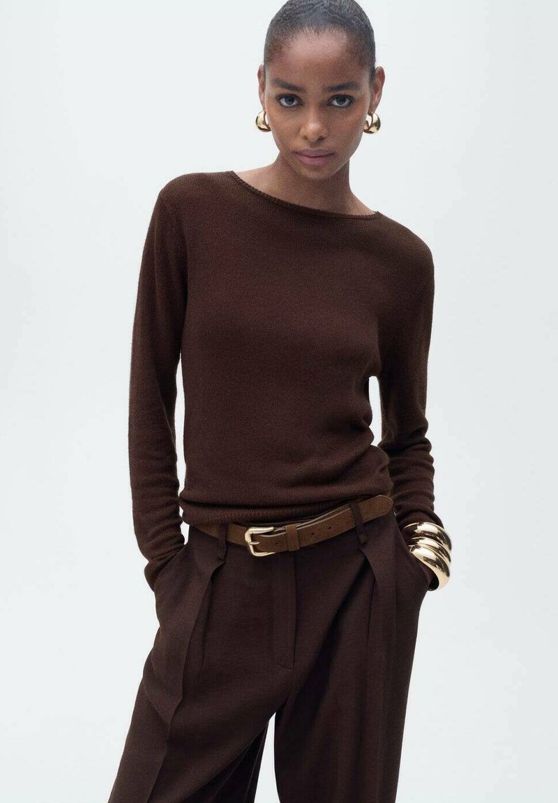 Pull en maille marron avec un col rond, associé à un pantalon marron taille haute et ajusté, et une ceinture dorée. Gros bracelet jonc en or en accent.