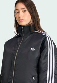 Une jeune femme aux cheveux longs porte une veste noire avec un col haut et des rayures blanches Adidas le long des manches, avec un logo sur la poitrine.