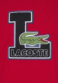 Tessuto rosso con una grande "L" nera e bianca in rilievo e "LACOSTE" sotto, e un design di coccodrillo ricamato in verde.
