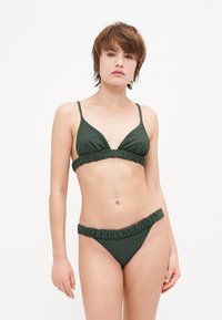 Tumši zaļa bikini komplekts ar fakturētu audumu. Augšdaļai ir trīsstūra forma un regulējamās siksnas; apakšdaļai ir volānu josta.