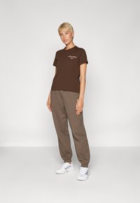 T-shirt in cotone marrone con maniche corte e logo bianco, abbinata a pantaloni da tuta marroni larghi. Scarpe bianche con strisce grigie completano l'outfit.
