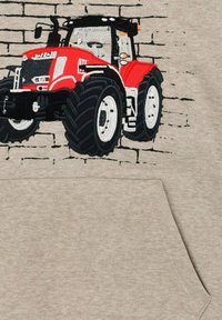 Grauer Pullover mit einem gestickten roten Traktor mit großen schwarzen Rädern, vor einem Backsteinhintergrund.