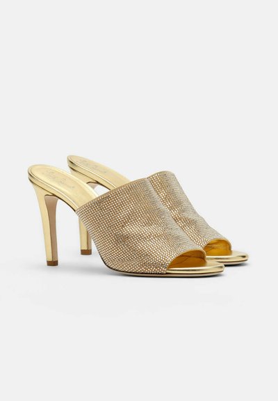 Mules à talons hauts ouverts dorés avec des brides larges ornées de strass et des talons fins sur un fond blanc uni.
