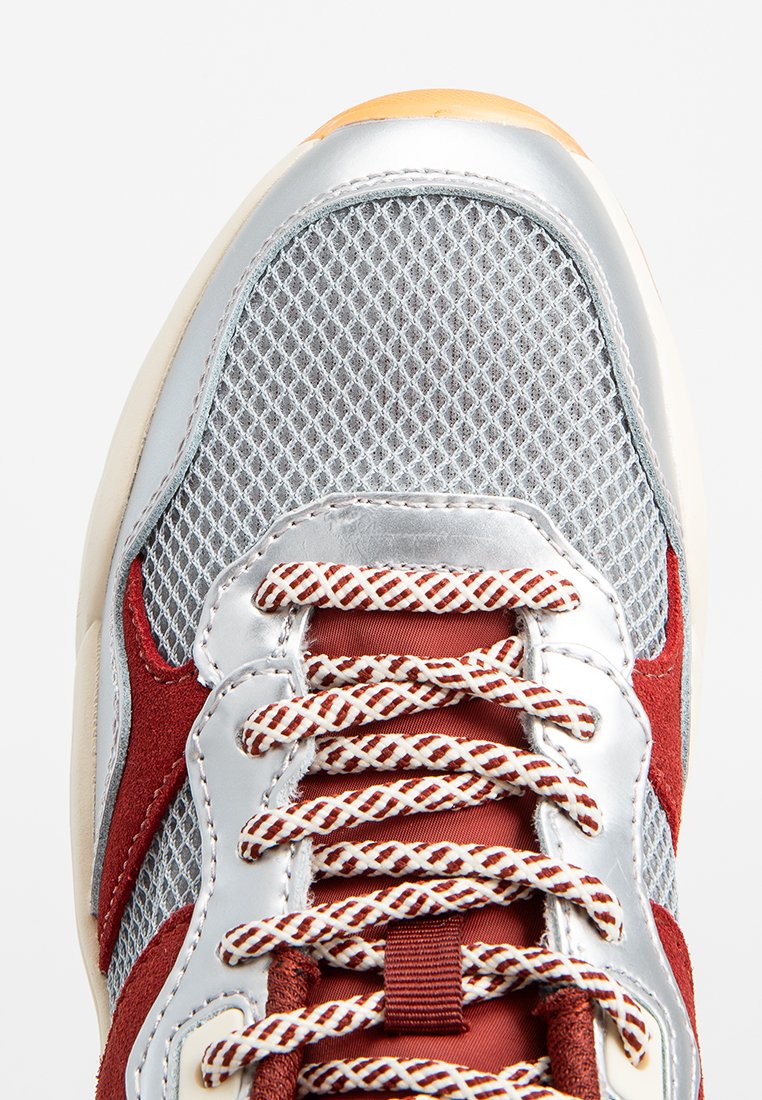 Grijze mesh sneaker met rode suède accenten, zilveren details en kruislings rode en witte veters. Afgeronde neus en gevoerde zool.