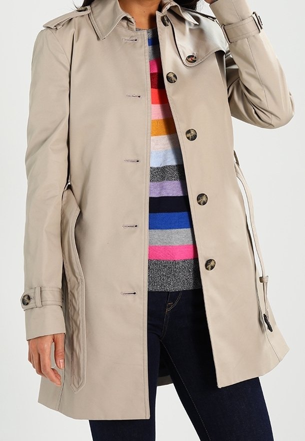 Beżowy trencz noszony rozpięty na wielokolorowym swetrze w paski i ciemnych dżinsach, z jedną ręką dotykającą głowy.