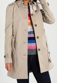 Vaaleanbeige trenchcoat avoinna monivärisen raidallisen neuleen ja tummien farkkujen päällä, yksi käsi pään koskettamassa.