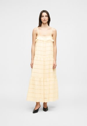 Kobieta ubrana w bezrękawową, jasnżółtą, warstwową sukienkę maxi z falbanami, czarne szpiczaste szpilki, stoi na jednolitym tle.