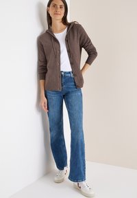 Braunem Cardigan mit Kapuze, Knöpfen und gerippten Bündchen, kombiniert mit blauen, ausgestellten Jeans und weißen Turnschuhen mit burgunderfarbenen Akzenten.