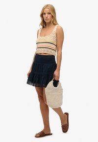 Häkel-Crop-Top mit mehrfarbigen Streifen, kombiniert mit einem marineblauen, gestuften Rock und einer gewebten beigefarbenen Handtasche. Braune flache Sandalen runden das Outfit ab.