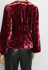 Top in velluto rosso con fiori, maniche lunghe trasparenti, busto attillato e dettaglio arricciato sulla schiena. Presenta uno sfondo nero con accenti testurizzati.