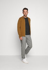 Cardigan cu fermoar bej, tricou negru, pantaloni cu model gri și adidași albi. Textură netedă, croială ajustată și stil casual.