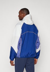 Veste coupe-vent avec capuche, présentant un design bicolore en bleu et blanc, fabriquée dans un tissu texturé avec des détails de surpiqûres contrastantes.