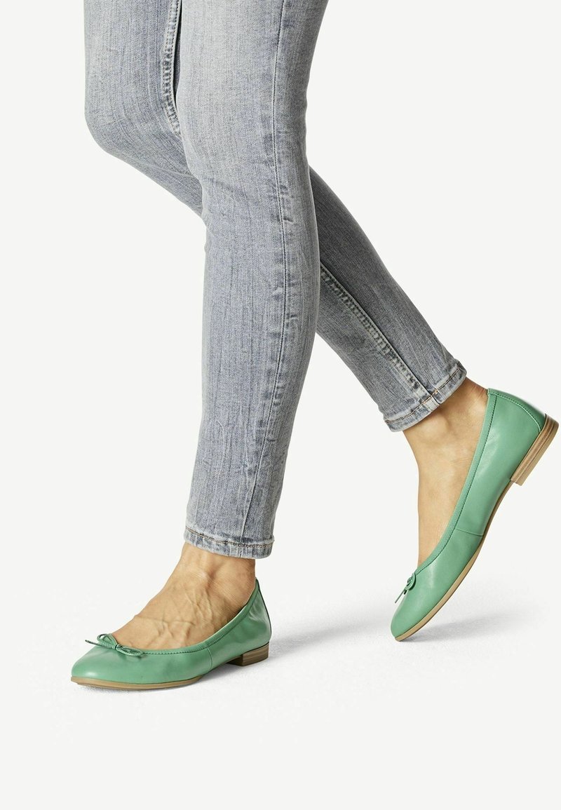 Ballet flats in pelle verde con tacco piatto, caratterizzate da un piccolo fiocco sulla punta, indossate con jeans skinny blu chiaro.