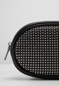 Schwarze Kosmetiktasche mit silberner Nietenverzierung, ovaler Form, glattem Lederfinish und einem Reißverschluss mit einem metallicfarbigen Zipperei.