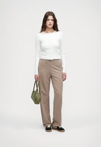 Biała bluzka z długim rękawem, zapinana na guziki, z falbaniastymi brzegami, w zestawie z beżowymi, luźnymi spodniami. Trzyma małą zieloną torebkę. Czarne buty.