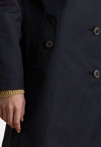 Abrigo de marina con un diseño de doble botonadura, dos botones y un bolsillo lateral. Los puños tienen un acento dorado de punto para añadir textura.