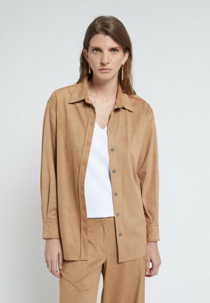 Donna con capelli castani lunghi che indossa una camicia beige con bottoni e pantaloni abbinati sopra una maglietta bianca, in piedi davanti a uno sfondo semplice.