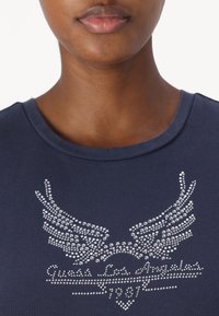 T-shirt blu navy a maniche corte, con un design di ali in strass argento e la scritta "Guess Los Angeles 1981" sulla parte anteriore. Texture liscia.