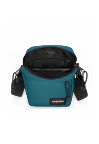 Tyrkysová látková kabelka přes rameno se zipem nahoře, černým vnitřkem, nastavitelným popruhem a logem na přední straně. Disponuje vnitřními kapsami.