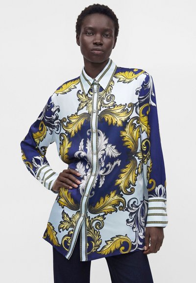 Personne aux cheveux courts portant une chemise à manches longues avec des motifs baroques ornés bleus, jaunes et blancs, debout avec une main sur la hanche.