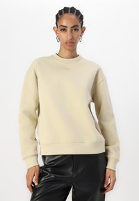 Sudadera beige de cuello redondo con un corte relajado, puños y dobladillo acanalados. Branding sutil en el pecho. Combinada con pantalones de piel sintética negros.