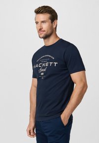 Hombre con barba y cabello corto, vistiendo una camiseta deportiva Hackett azul marino y pantalones a juego, de pie con una mano en el bolsillo.