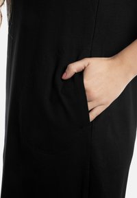 Zwarte jurk met een gestructureerd oppervlak en een ronde zak. Een hand is in de zak geplaatst, wat het functionele ontwerp van de jurk benadrukt.