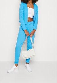 Nebeský modrý oblek s přizpůsobeným sakem a odpovídajícími kalhotami. Bílé crop top pod ním, doplněný bílou texturovanou taškou na rameno a bílými teniskami.