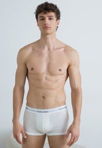 Bijele pamučne bokserice s elastikašem s logotipom Calvin Klein, s prilagođenim dizajnom i glatkom teksturom. Model stoji uz jednostavnu pozadinu.