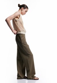 Beige mesh tanktop med lyse kanter, sammen med højtaljede olivengrønne bukser med vide ben og sorte sandaler, vist i profil.