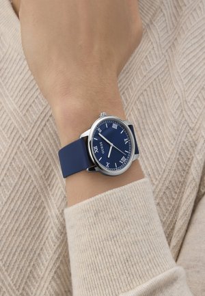 Mornarsko modra ura z okroglo srebrno ohišje, rimski številčnice in gladkim gumijastim trakom, nosi na svetlem pletenem puloverju.